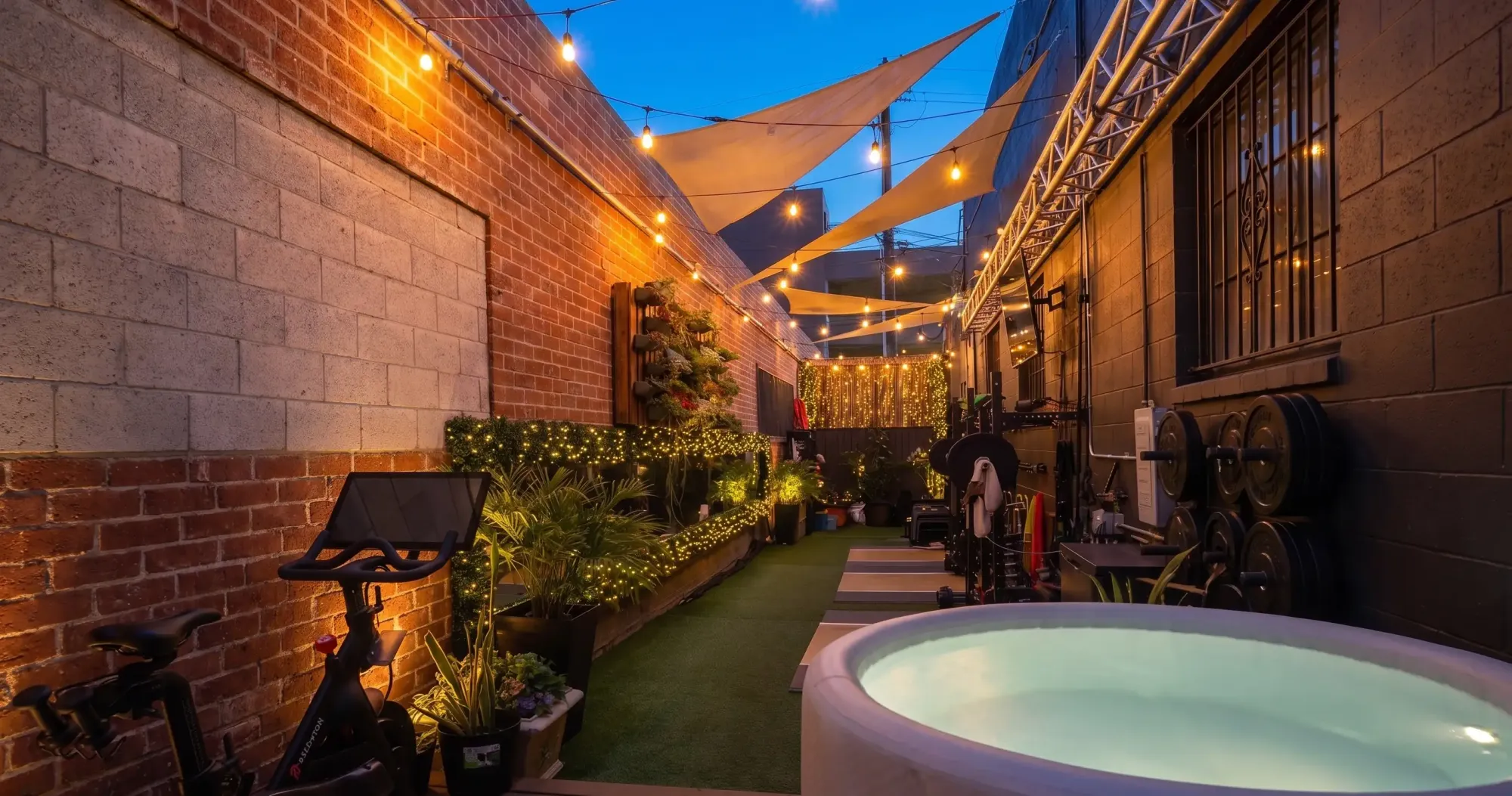 The courtyard at The Recording Club at dusk: string lights, shade sails, planters and a lighting truss along the brick walls